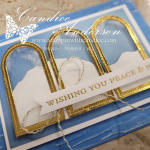 A close-up of a minimalist Christmas card design featuring three arched gold windows, layered with white and blue elements. The card includes a sentiment 'Wishing You Peace & Joy' displayed on a banner, with delicate twine accents.