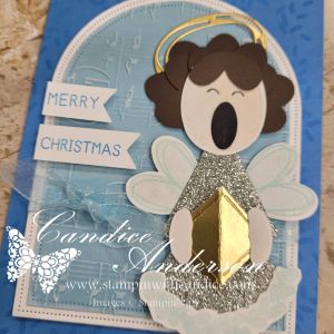 A whimsical holiday card featuring a punched angel with curly hair, shimmering wings, and a golden gift. The card includes 'Merry' and 'Christmas' sentiments on banners, with a blue background and decorative elements.