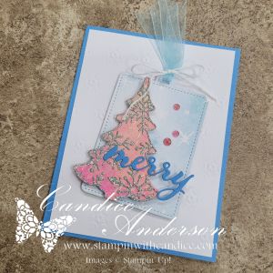 A handmade card featuring a decorated tag with a pink and gold Christmas tree and the word 'merry,' placed on a blue cardstock background.