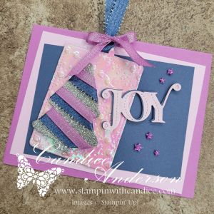 A colorful card featuring the word 'JOY' in white, embellished with sparkly ribbons and layered iridescent elements in shades of pink and purple.