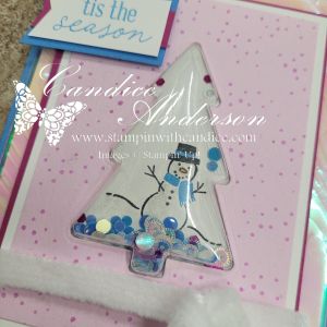 A festive shaker card featuring a clear tree shape filled with sequins and a snowman drawing, set against a pink polka dot background with a sentiment that says 'tis the season.'