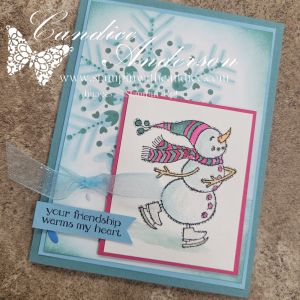 A handmade card featuring a whimsical snowman skating on ice, surrounded by a soft, snowy background with blue hues. The snowman is wearing a colorful scarf and a hat, and there is a sentiment that reads, 'your friendship warms my heart.' The card is adorned with a sheer ribbon and decorative elements inspired by a playful woodland theme.