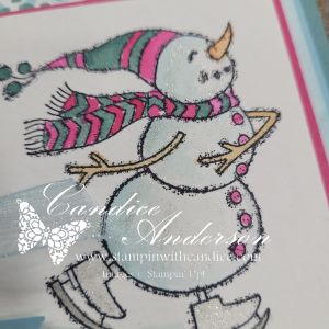 A whimsical snowman with a pink and teal scarf and a carrot nose, smiling while skating. The image is outlined with glitter for added shimmer.
