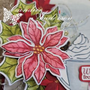 Close-up view of a handcrafted card featuring a pink poinsettia flower, green leaves, and a cozy mug of cocoa with whipped topping.