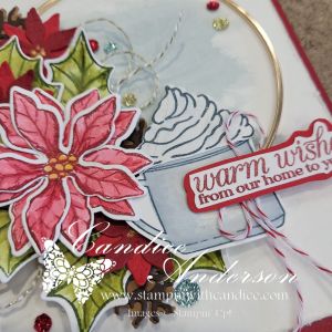 Close-up of a festive winter card featuring a watercolored poinsettia, a mug of cocoa topped with whipped cream, and a decorative sentiment that reads 'warm wishes from our home to yours,' arranged around a gold hoop.