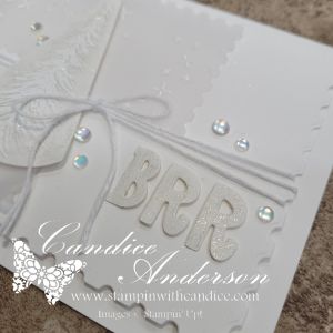 A close-up of a handmade card featuring layered white elements, including the letters 'BRR' embellished with sparkle, tied with white twine, and adorned with iridescent dots for a snow-kissed effect.