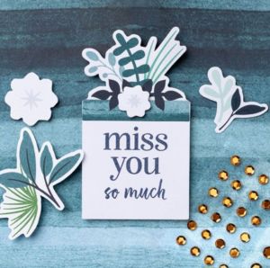 A card featuring the message 'miss you so much' surrounded by decorative paper cutouts of leaves and flowers, with gold sequins on a textured blue background.