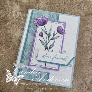 A handmade floral greeting card featuring three purple flowers with green leaves, layered on a soft blue background and bordered by a light purple frame. The card includes a white rectangular label with the text 'dear friend' on it.