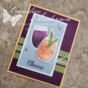A handmade card featuring two stylized drink glasses, one purple and one orange, with a sprig of greenery, set against a textured, dark purple background and accented with a 'Cheers' message and ribbon.