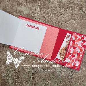 An open greeting card featuring the phrase 'I ADORE YOU' on one page, with a decorative pattern of hearts and a $10 gift card inserted on the side.