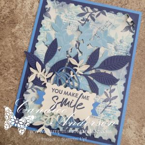 A handcrafted card featuring layers of blue paper with floral and leaf embellishments, and the message 'YOU MAKE ME SMILE' in the center.