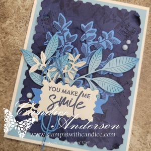 A handmade card featuring blue and white paper leaves and flowers, with the message 'YOU MAKE ME SMILE' prominently displayed on a scalloped tag. The background is a textured dark blue, enhancing the layered design.