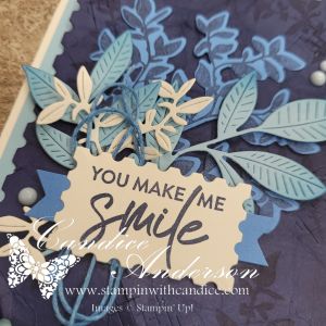 A handmade card featuring a blue and purple floral design, with layered leaves and a tag that reads 'YOU MAKE ME SMILE'.
