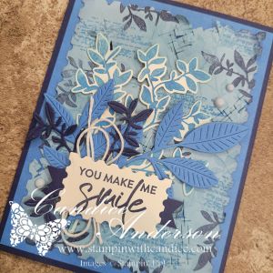 A handmade card featuring a blue background with a layered design, adorned with various leaf shapes in different shades of blue. The message 'You Make Me Smile' is prominently displayed in a decorative frame at the center.