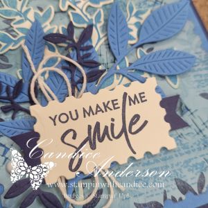 A close-up of a handmade card featuring a decorative tag that reads 'YOU MAKE ME SMILE', surrounded by various shades of blue leaves and twine.