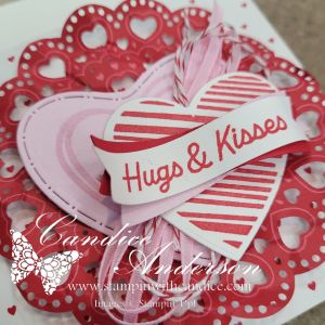 A close-up view of a decorative Valentine's Day card featuring layered hearts in shades of pink and red, with a ribbon that reads 'Hugs & Kisses'. The background includes a lace-like doily pattern.