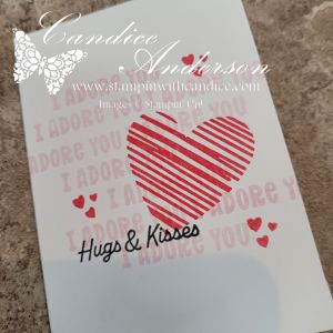 A greeting card design featuring a large striped heart, the words 'I adore you' repeated in the background, and 'Hugs & Kisses' written prominently at the bottom.