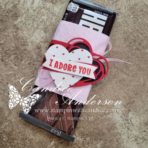 A wrapped chocolate bar decorated with a pink and red heart-shaped tag that says 'I ADORE YOU', resting on a textured surface.
