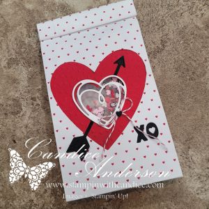 A decorative treat bag featuring a large red heart with an arrow, a transparent heart-shaped window filled with confetti, and a string bow. The background is white with small red hearts.