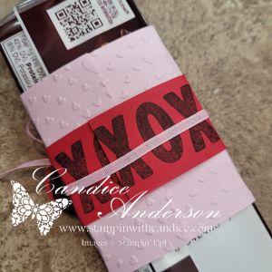 A wrapped chocolate bar with a pink embossed cover featuring small hearts, a red band displaying the word 'XOXO' in black, and a decorative ribbon.