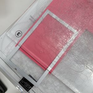 A close-up view of a metal cutting die positioned over a pink cardstock, with an embossed texture visible beneath the die. The setup appears to be on a craft table, including a clear base plate.
