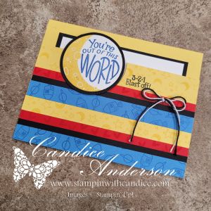 A colorful greeting card featuring the phrase 'You're out of this WORLD' in a circular design. The card has a yellow background with horizontal stripes of red, blue, and black, adorned with a bow made of black string.
